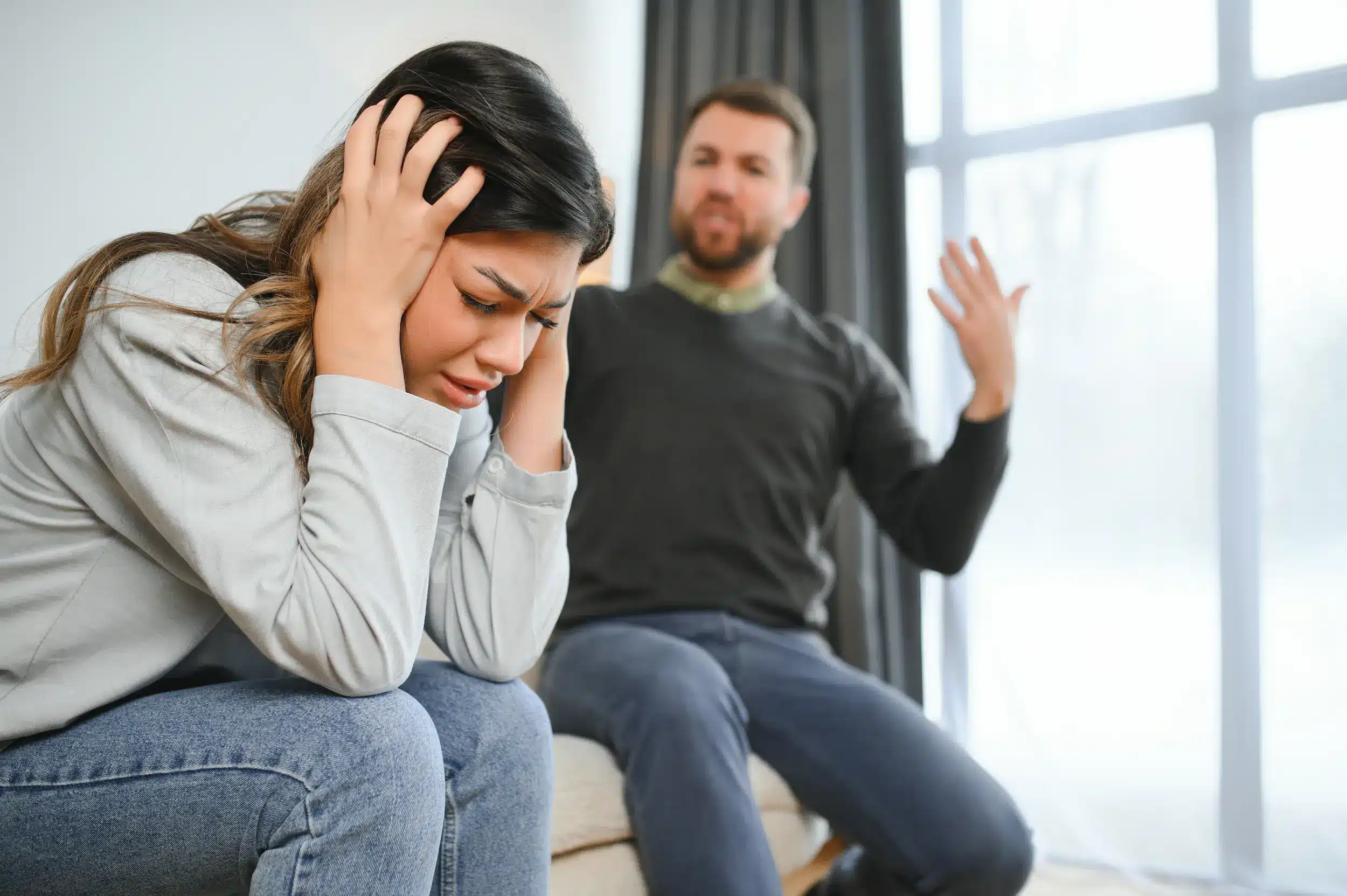 Mulher angustiada segurando a cabeça durante uma discussão, com o homem ao fundo, expressando raiva, ilustrando um cenário de manipulação emocional.
