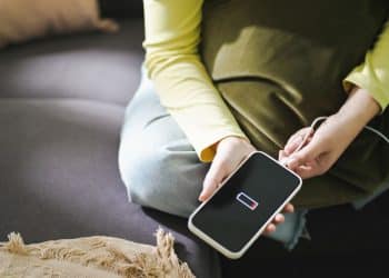Celular com bateria baixa sendo conectado ao carregador por uma pessoa sentada no sofá.