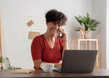 Uma mulher com cabelos pretos curtos e cacheados, vestindo uma blusa de tricô vermelha, sentada em uma mesa de madeira em um ambiente de escritório doméstico bem iluminado. Ela está sorrindo e falando ao celular, enquanto olha para a tela de um laptop cinza aberto. Sobre a mesa há uma xícara branca e um caderno. Ao fundo, vê-se uma estante de madeira e uma planta.