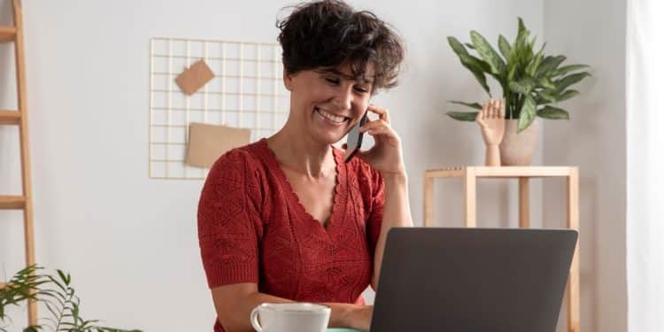 Uma mulher com cabelos pretos curtos e cacheados, vestindo uma blusa de tricô vermelha, sentada em uma mesa de madeira em um ambiente de escritório doméstico bem iluminado. Ela está sorrindo e falando ao celular, enquanto olha para a tela de um laptop cinza aberto. Sobre a mesa há uma xícara branca e um caderno. Ao fundo, vê-se uma estante de madeira e uma planta.