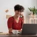 Uma mulher com cabelos pretos curtos e cacheados, vestindo uma blusa de tricô vermelha, sentada em uma mesa de madeira em um ambiente de escritório doméstico bem iluminado. Ela está sorrindo e falando ao celular, enquanto olha para a tela de um laptop cinza aberto. Sobre a mesa há uma xícara branca e um caderno. Ao fundo, vê-se uma estante de madeira e uma planta.