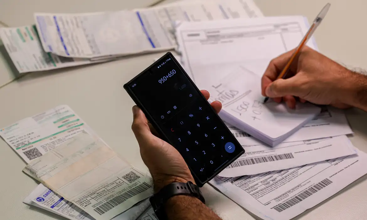 Empresário segurando celular com calculadora e fazendo contas em meio a contas e boletos representando o pagamento de impostos do Simples Nacional