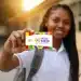 Jovem estudante negra, com cabelos longos trançados, uniforme escolar branco e mochila, sorrindo e exibindo o cartão colorido do programa Pé-de-Meia com a frase "A poupança do ensino médio", em frente a uma escola com luz solar ao fundo, representando os estudantes do ensino médio da rede pública que podem ser beneficiados pelo programa em 2026