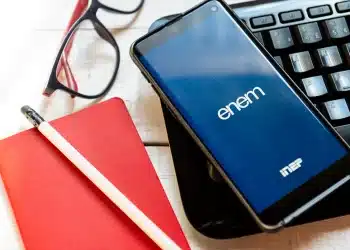 Imagem mostrando o celular com o logo do Enem 2026 na tela, com caderno, caneta e óculos sobre a mesa. A tela do celular exibe o aplicativo do Enem.