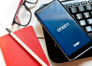 Celular com logo do Enem Inep ao lado de caderno vermelho e óculos, representando a isenção da taxa de inscrição do Enem 2026