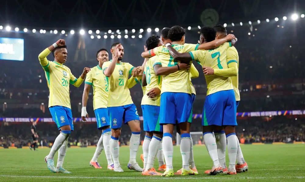 Jogadores da Seleção Brasileira de futebol, vestindo o uniforme oficial com camisa amarela e calção azul, reunidos em um abraço coletivo no centro do campo durante uma partida noturna em um estádio lotado.