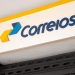 Placa branca com logo dos Correios em azul e amarelo na fachada da agência, com grade de ferro.