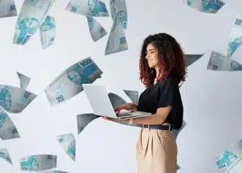 Mulher usando notebook enquanto cédulas de dinheiro aparecem ao redor, representando trabalho digital e geração de renda.