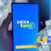 Tela do aplicativo CAIXA Tem em um smartphone, com a logo do serviço e cédulas de dinheiro ao fundo, relacionado ao processo de liberação de conta para estudantes menores de idade no programa Pé-de-Meia.