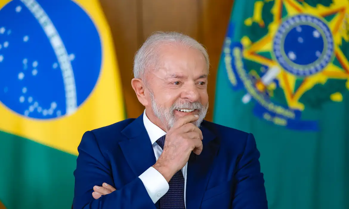 Presidente Lula sorrindo durante evento no Palácio do Planalto, com as bandeiras do Brasil e da República ao fundo, após anunciar envio do projeto de lei sobre a escala 6x1