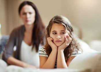 Foto em plano médio de uma menina de aproximadamente 10 anos sentada em um sofá, com as mãos apoiando o rosto e expressão de profundo desânimo e isolamento emocional. Ao fundo, de forma desfocada, uma mulher adulta observa a criança com uma expressão severa e crítica. A iluminação é natural e clara.