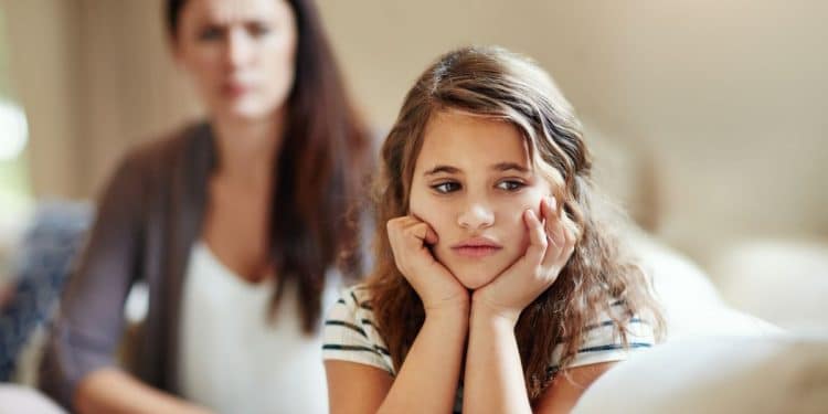 Foto em plano médio de uma menina de aproximadamente 10 anos sentada em um sofá, com as mãos apoiando o rosto e expressão de profundo desânimo e isolamento emocional. Ao fundo, de forma desfocada, uma mulher adulta observa a criança com uma expressão severa e crítica. A iluminação é natural e clara.