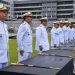 Plano médio de uma cerimônia militar oficial da Marinha do Brasil. Fileiras de Oficiais da Marinha, em pé e em continência, vestindo o uniforme branco de gala com insígnias douradas e quepe. Eles estão perfilados sobre um gramado verde, olhando para a direita. No centro da imagem, sobre uma longa mesa coberta por um pano preto, estão várias espadas cerimoniais com cabos e bainhas douradas, prontas para serem entregues. Ao fundo, vê-se um grande edifício militar cinza e branco e várias bandeiras hasteadas. No canto inferior direito, há uma pequena marca d'água com o símbolo da Marinha. A luz é natural e de um dia nublado.