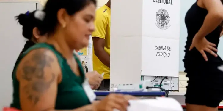 Mesária e eleitores em cabine de votação durante as eleições, destacando os mesários no processo eleitoral.