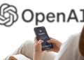 Pessoa segurando iPhone com tela de boas-vindas do ChatGPT, logo da OpenAI ao fundo em parede branca