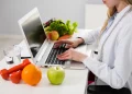 Estudante de Nutrição analisando alimentos saudáveis enquanto utiliza notebook em ambiente de estudo.
