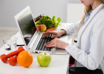 Estudante de Nutrição analisando alimentos saudáveis enquanto utiliza notebook em ambiente de estudo.