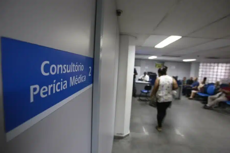 Sala de atendimento de perícia médica do INSS, com sinalização para o consultório e pessoas aguardando.