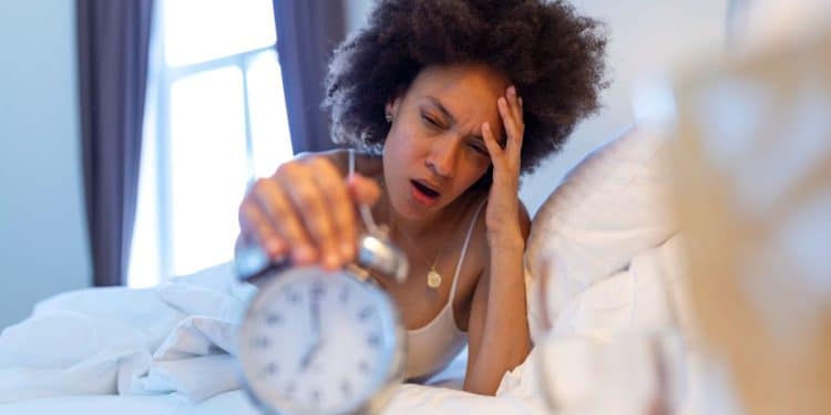 Close-up de uma mulher negra com cabelos cacheados, deitada em uma cama com lençóis brancos. Ela está com uma expressão de exaustão, bocejando e com a mão na testa, enquanto estica o outro braço para tentar desligar um despertador analógico prata em primeiro plano. O despertador está levemente desfocado. Ao fundo, uma janela com cortinas escuras e a luz da manhã entrando.