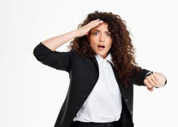 Mulher com cabelo cacheado e traje formal, olhando ansiosa para o relógio, refletindo sobre os motivos psicológicos dos atrasos.
