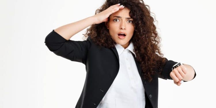 Mulher com cabelo cacheado e traje formal, olhando ansiosa para o relógio, refletindo sobre os motivos psicológicos dos atrasos.