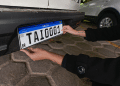 Mãos com tatuagens posicionando placa Mercosul branca com faixa azul e código TAI0001 em para-choque traseiro de carro preto