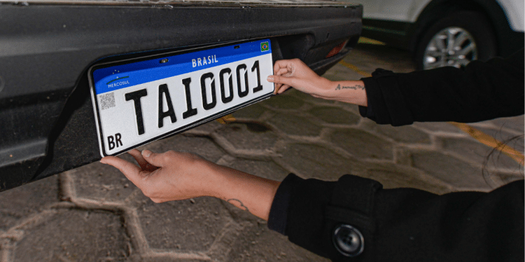 Mãos com tatuagens posicionando placa Mercosul branca com faixa azul e código TAI0001 em para-choque traseiro de carro preto