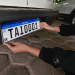 Mãos com tatuagens posicionando placa Mercosul branca com faixa azul e código TAI0001 em para-choque traseiro de carro preto