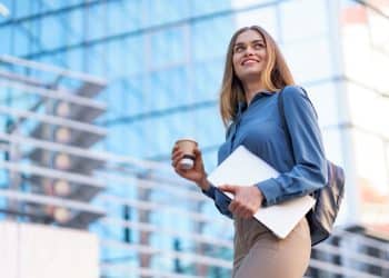 Mulher jovem e sorridente com cabelo loiro, usando camisa social azul e calça bege, segura um copo de café e um notebook enquanto olha para o alto na frente de um prédio espelhado de escritórios.