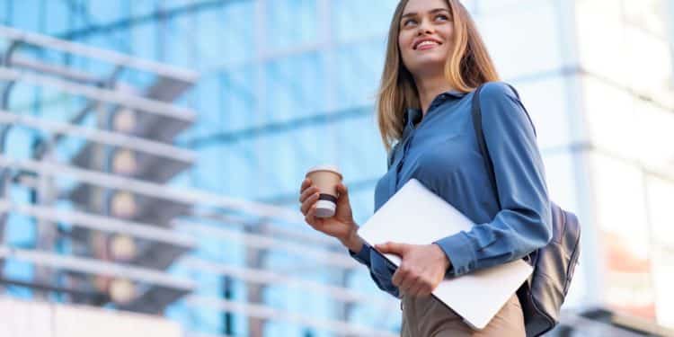 Mulher jovem e sorridente com cabelo loiro, usando camisa social azul e calça bege, segura um copo de café e um notebook enquanto olha para o alto na frente de um prédio espelhado de escritórios.
