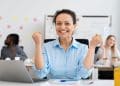 Mulher sorridente de camisa azul comemora com os punhos cerrados, sentada à mesa com notebook e colegas ao fundo, simbolizando as profissões em alta para 2026.