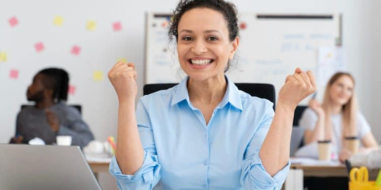 Mulher sorridente de camisa azul comemora com os punhos cerrados, sentada à mesa com notebook e colegas ao fundo, simbolizando as profissões em alta para 2026.