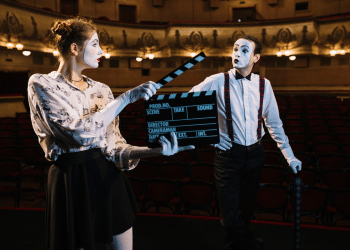 Mímicos com maquiagem branca e suspensórios vermelhos posam com claquete de cinema em teatro vazio.