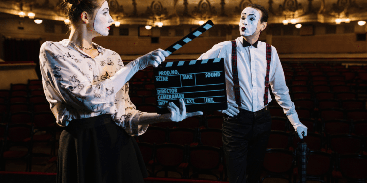 Mímicos com maquiagem branca e suspensórios vermelhos posam com claquete de cinema em teatro vazio.