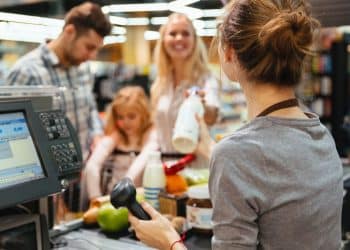 Vendedora de supermercado interagindo com clientes durante o pagamento, em uma loja movimentada.