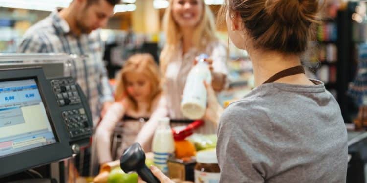 Vendedora de supermercado interagindo com clientes durante o pagamento, em uma loja movimentada.