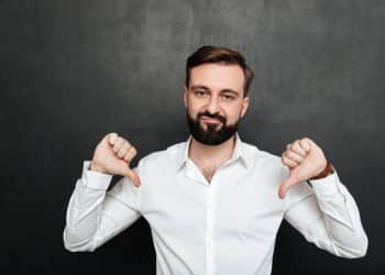 Homem de camisa branca fazendo gesto de polegar para baixo em fundo escuro, simbolizando frustração com baixos salários.