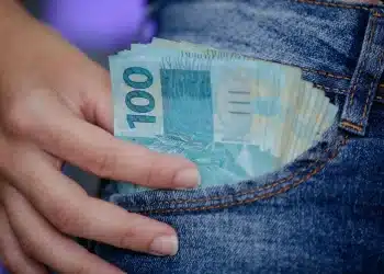 Notas de 100 reais sendo retiradas do bolso de um jeans, simbolizando o alcance do salário ideal no Brasil por meio de profissões técnicas.