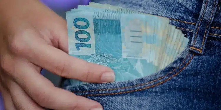 Notas de 100 reais sendo retiradas do bolso de um jeans, simbolizando o alcance do salário ideal no Brasil por meio de profissões técnicas.