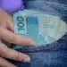 Notas de 100 reais sendo retiradas do bolso de um jeans, simbolizando o alcance do salário ideal no Brasil por meio de profissões técnicas.