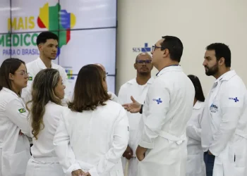 Grupo de médicos com jalecos brancos do SUS conversando em frente ao painel do programa Mais Médicos para o Brasil, representando os profissionais selecionados para atuar em regiões prioritárias do SUS