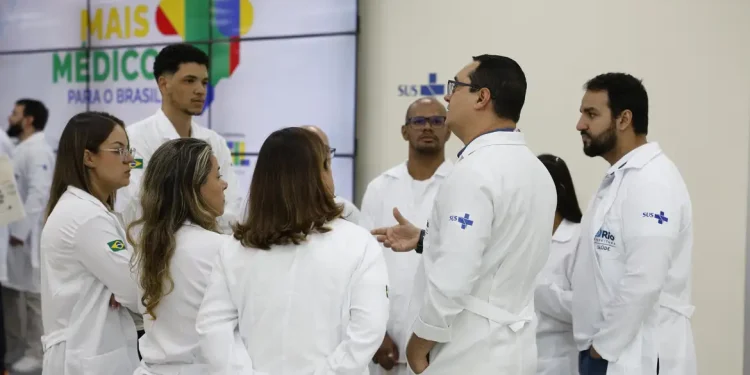 Grupo de médicos com jalecos brancos do SUS conversando em frente ao painel do programa Mais Médicos para o Brasil, representando os profissionais selecionados para atuar em regiões prioritárias do SUS