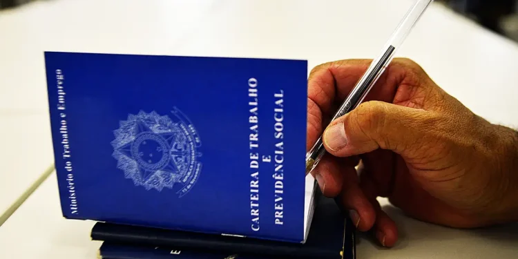 Mão segurando caneta sobre carteiras de trabalho azuis em ilustração sobre as mudanças propostas na CLT para o direito à desconexão do trabalhador