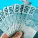 Mão segurando várias notas de 100 reais em leque representando o reajuste do salário mínimo em 2027 para aposentados e pensionistas do INSS