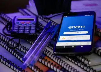Estudante se preparando para o Enem com celular aberto no aplicativo, cadernos, canetas e calculadora sobre a mesa.
