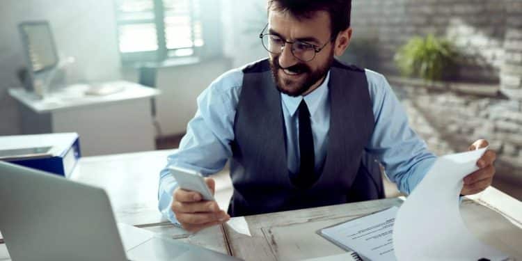 Profissional analisa documentos e utiliza celular em ambiente de escritório corporativo.