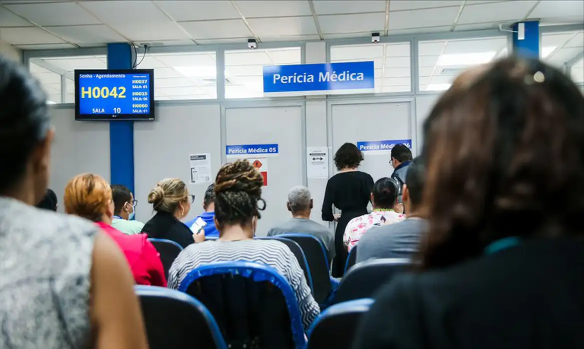 Pessoas aguardam atendimento em sala de espera da Perícia Médica do INSS, com painel eletrônico de senhas exibindo a chamada H0042 e placas indicativas das salas