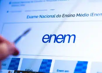 Mão segurando caneta em frente à tela de computador exibindo o site oficial do Enem, representando a preparação para a redação do Exame Nacional do Ensino Médio.