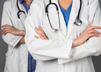 Foto em plano médio e em close, sem mostrar o rosto, de dois profissionais de medicina em pé, um ligeiramente atrás do outro. Ambos usam jalecos brancos sobre roupas hospitalares azuis (um azul royal e o outro azul claro). Eles estão com os braços cruzados na altura do peito e têm estetoscópios pendurados no pescoço. O fundo é cinza uniforme e desfocado.