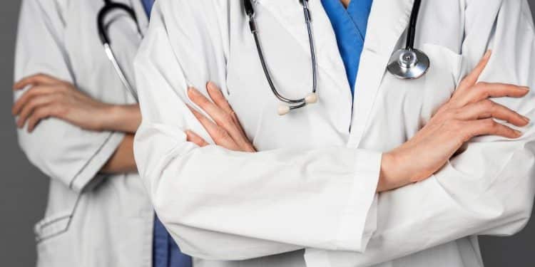 Foto em plano médio e em close, sem mostrar o rosto, de dois profissionais de medicina em pé, um ligeiramente atrás do outro. Ambos usam jalecos brancos sobre roupas hospitalares azuis (um azul royal e o outro azul claro). Eles estão com os braços cruzados na altura do peito e têm estetoscópios pendurados no pescoço. O fundo é cinza uniforme e desfocado.